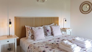 Un dormitorio con cabecera de madera, cama con ropa de cama floral, dos mesitas de noche con lámparas y un espejo redondo en la pared.