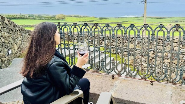 Una persona sentada en un banco, sosteniendo una copa de vino tinto, con un muro de piedra y una cerca de metal al fondo.