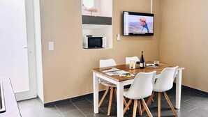 Un comedor con una mesa de madera, sillas blancas, un televisor montado en la pared y un microondas en un estante.