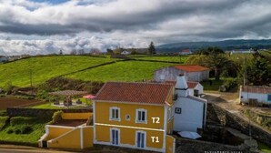 Paisaje rural con un edificio amarillo marcado T1 y T2, rodeado de campos verdes y un cielo nublado.