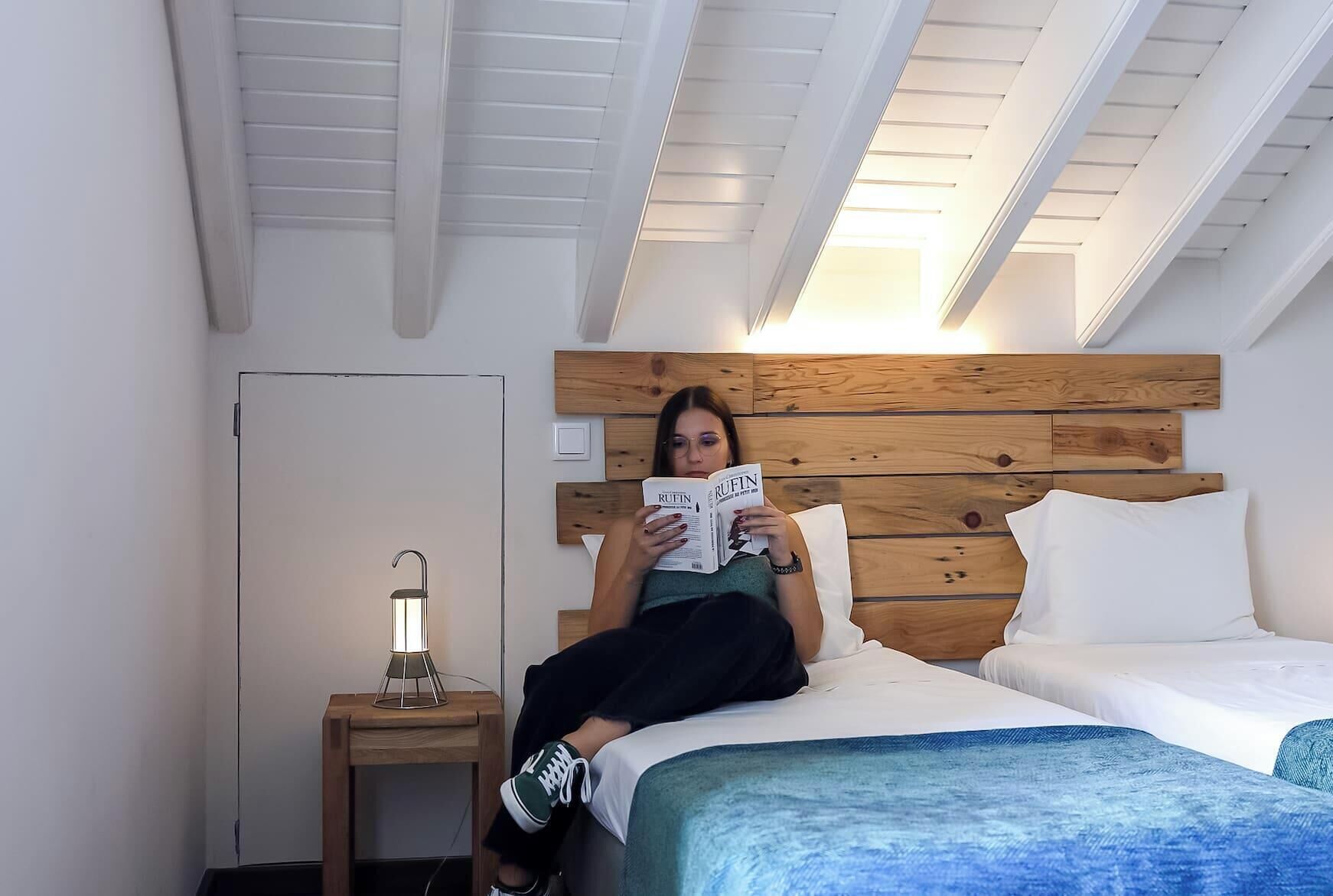 A person reading a magazine on a bed with a wooden headboard, a bedside table with a lamp, and a window with skylights.