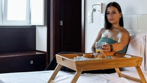 Eine Person sitzt auf einem Bett, auf einem Tablett befinden sich Speisen und ein Glas Orangensaft.