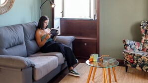 Eine Person sitzt auf einem grauen Sofa und liest ein Buch, in der Nähe steht ein Glastisch und ein blumiger Sessel.