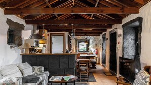 Un interior rústico con vigas de madera a la vista, chimenea de piedra, un sofá y una zona de cocina.