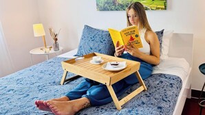 A person sitting on a bed with a tray of food, reading a book, in a room with a painting on the wall. - Casa da Maresia - By Ponta Negra Azores (Praia da Vitória)