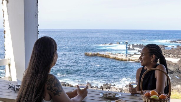 Two people seated at a wooden table with a view of the ocean, a rocky pier, and a lighthouse in the background. - Casa da Maresia - By Ponta Negra Azores (Praia da Vitória)