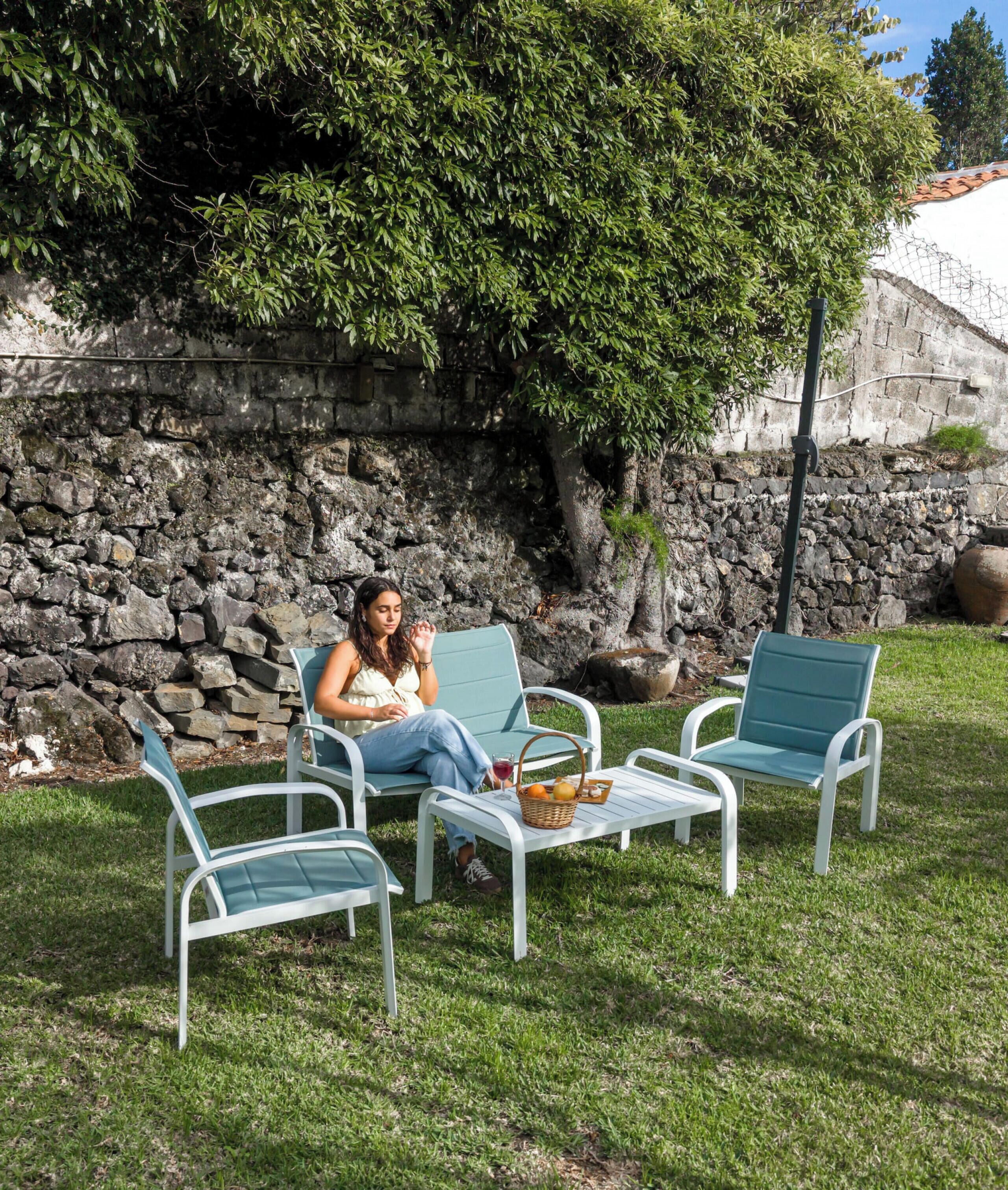 Una persona sentada en una silla en un área exterior con césped, un muro de piedra y un árbol al fondo.