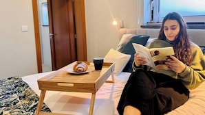 A person sitting on a bed reading a book, with a wooden side table in front of them holding a croissant and a cup.