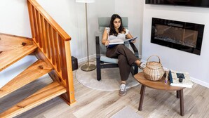 A person sitting on a chair reading a book in a room with a wooden staircase, a floor lamp, and a flat-screen TV mounted on the wall.