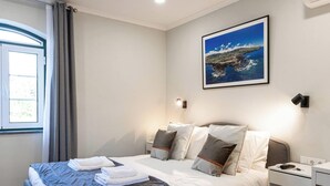 A hotel room with a bed, a nightstand, a wall-mounted light, a window with curtains, and a framed picture of a coastal landscape.