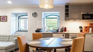 A modern kitchen with a dining area, featuring a round table with chairs, a pendant light, and a view through the windows.