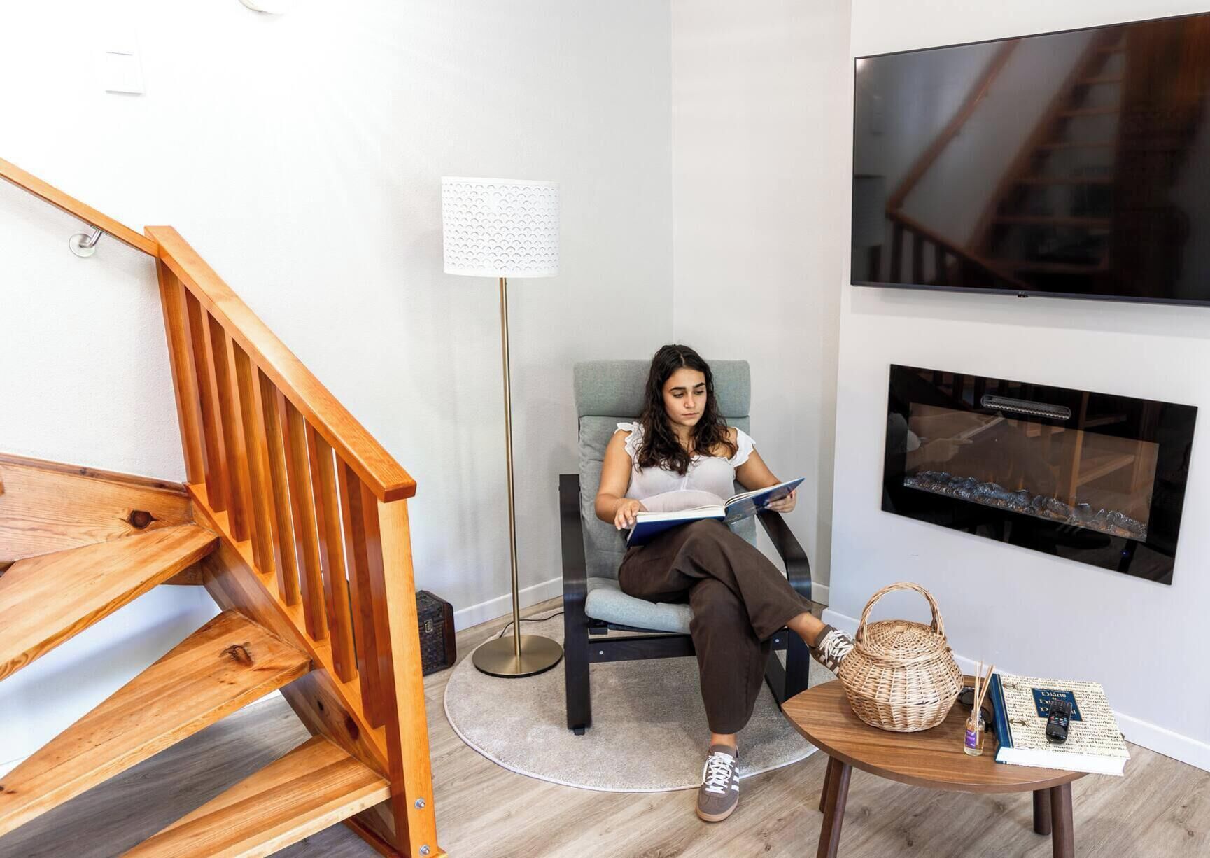 Una persona asseguda en una cadira llegint un llibre en una habitació amb una escala de fusta, una làmpada de peu i un televisor de pantalla plana penjat a la paret.