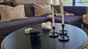 Ein modernes Wohnzimmer mit einem dunklen Sofa, gemusterten Kissen, einem runden Couchtisch und zwei hohen Kerzen.
