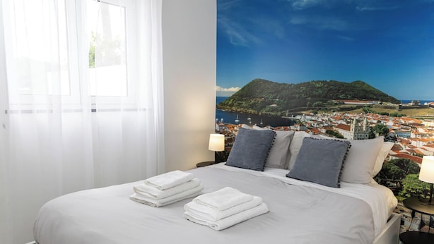 Una camera d'albergo con un letto, tende bianche, vista su una cittadina costiera e una lampada sul comodino.