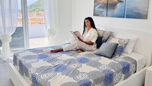 Eine Person sitzt auf einem Bett und liest ein Buch in einem Raum mit einem großen Fenster, einem Gemälde und Blick auf eine Landschaft.