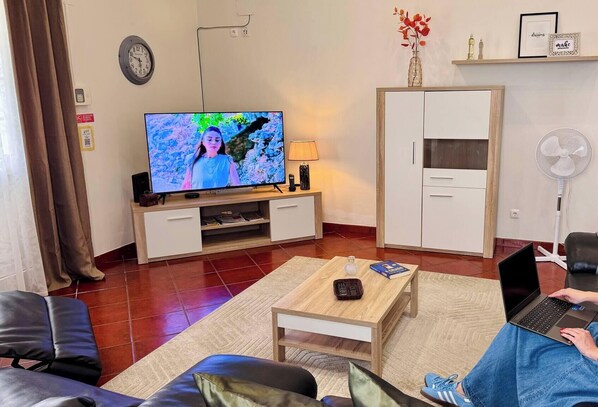 Un salon avec un téléviseur, un ordinateur portable posé sur une table, un canapé, un meuble et un ventilateur.