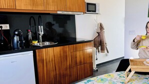 A compact kitchen with a microwave, coffee maker, and sink. There is a bed with a floral bedspread and a wooden table with a cup and saucer. - Recanto das 5 T0 by PontaNegraAzores (Angra do Heroísmo)