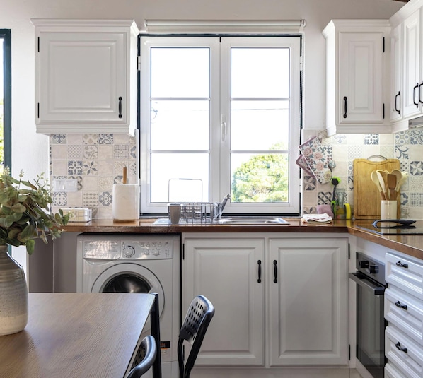 A kitchen with white cabinets, a washing machine, a dining table, and a window with a view of greenery.