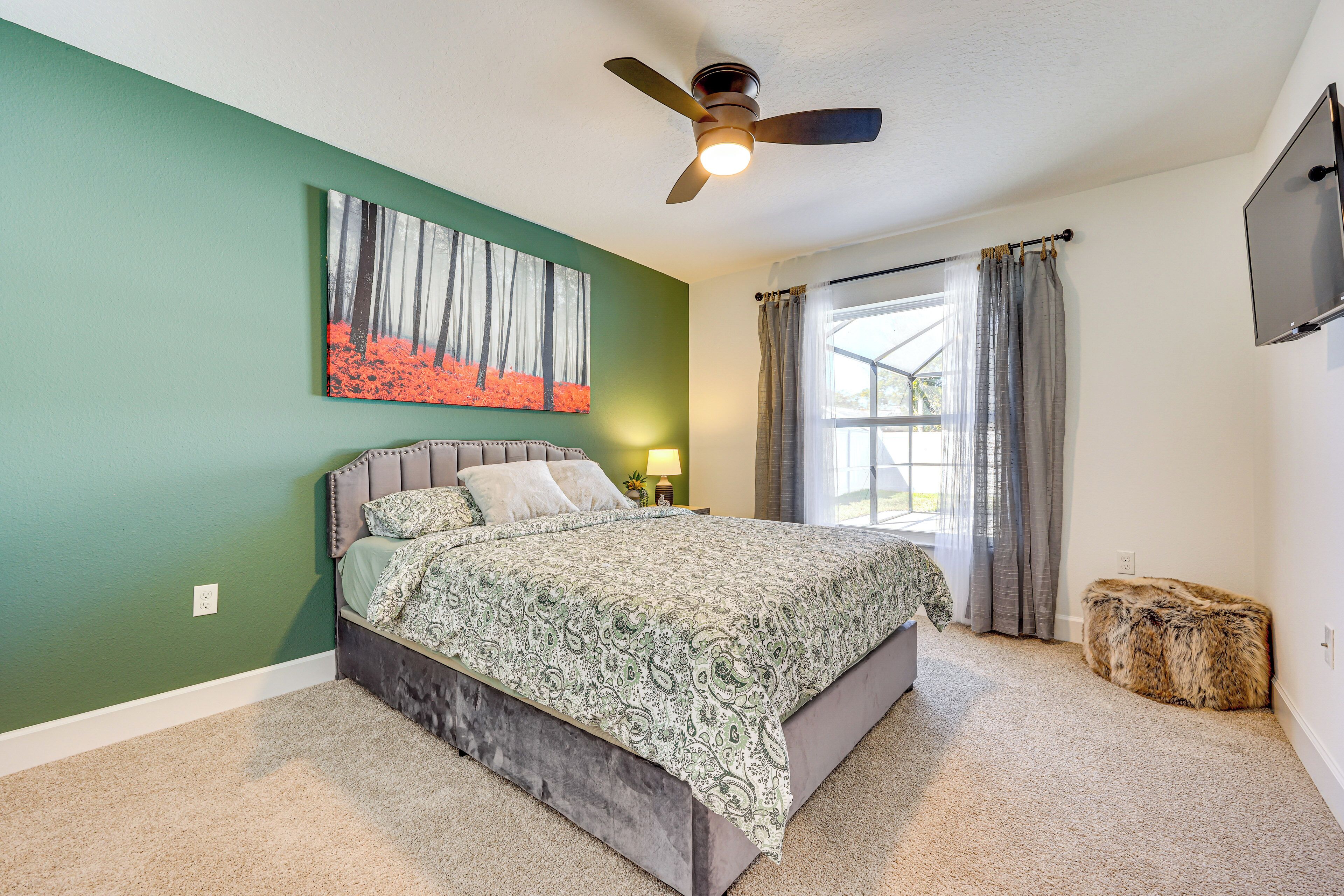 A bedroom with a bed, a ceiling fan, a painting, a window with curtains, and a television on the wall.