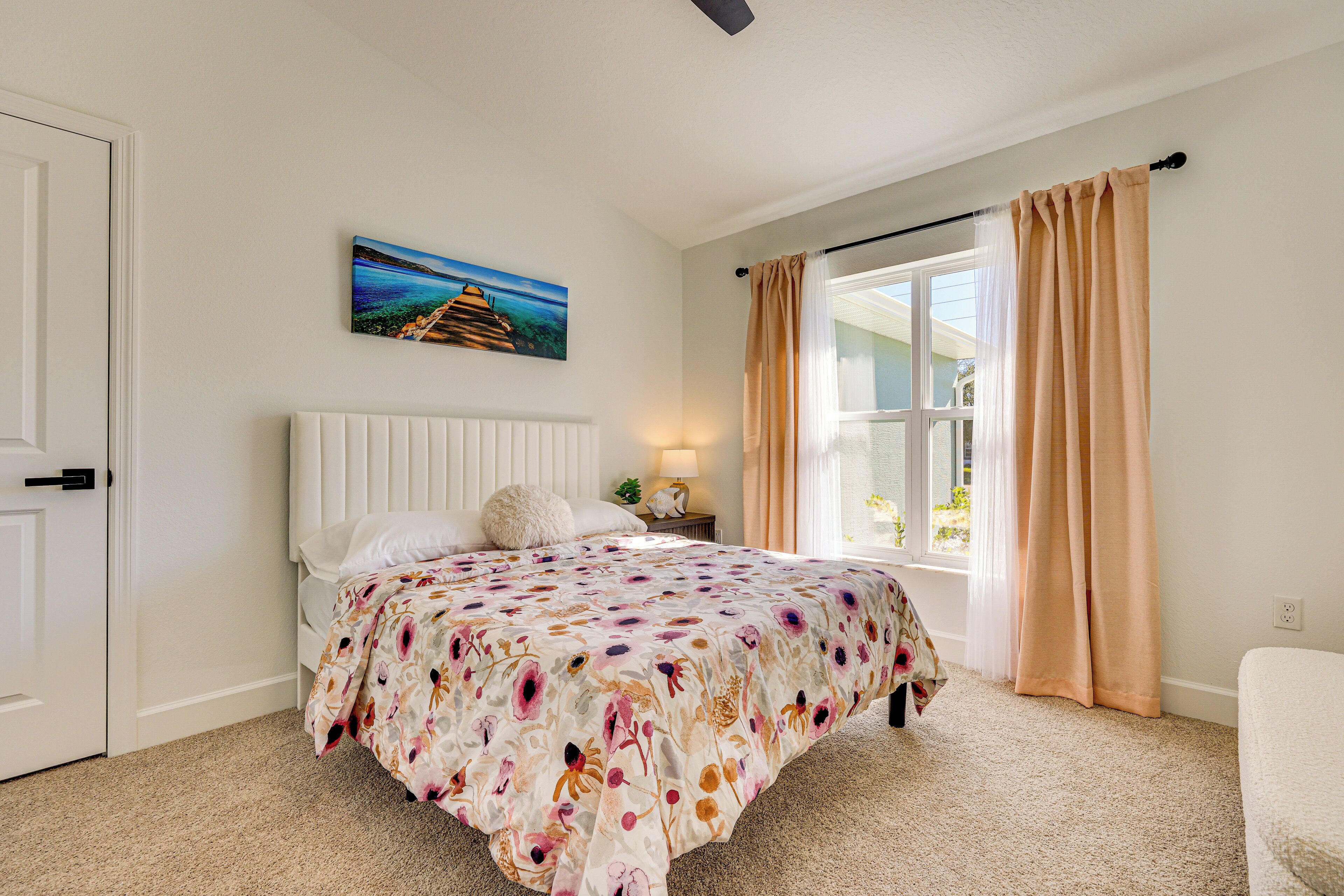 A bedroom with a bed, a nightstand, a lamp, a window with curtains, and a painting on the wall.