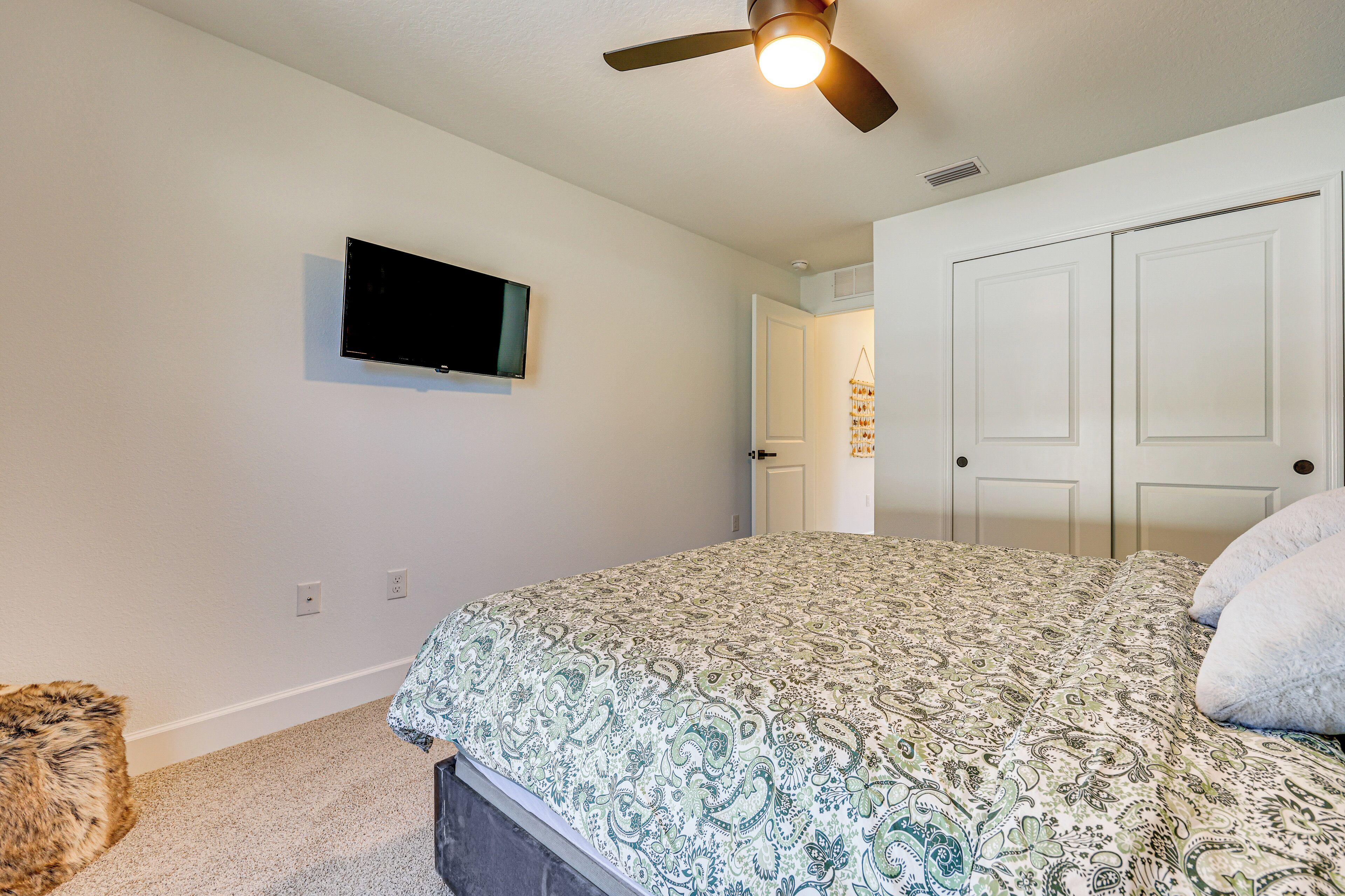 A bedroom with a bed, a ceiling fan, a mounted TV, and a door leading to another room.