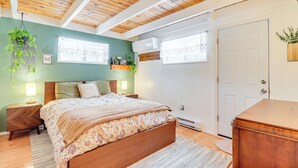 Ein Schlafzimmer mit Holzbalkendecke, einem Bett mit blumigem Überwurf, zwei Nachttischen mit Lampen, einem Fenster mit Spitzenvorhängen und einer Tür.