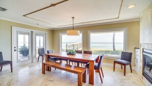 Uma ampla área de jantar com mesa e bancos de madeira, grandes janelas com vista para o oceano e uma lareira à direita.
