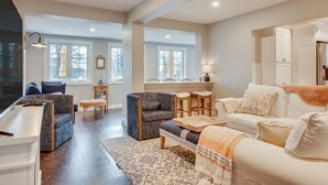 A spacious living room with a sofa, armchairs, a coffee table, and a TV. There are windows with curtains, a fireplace, and a kitchen area visible. - 1 Mi to Drakes Island Beach: Cottage w/ Deck! (Wells)