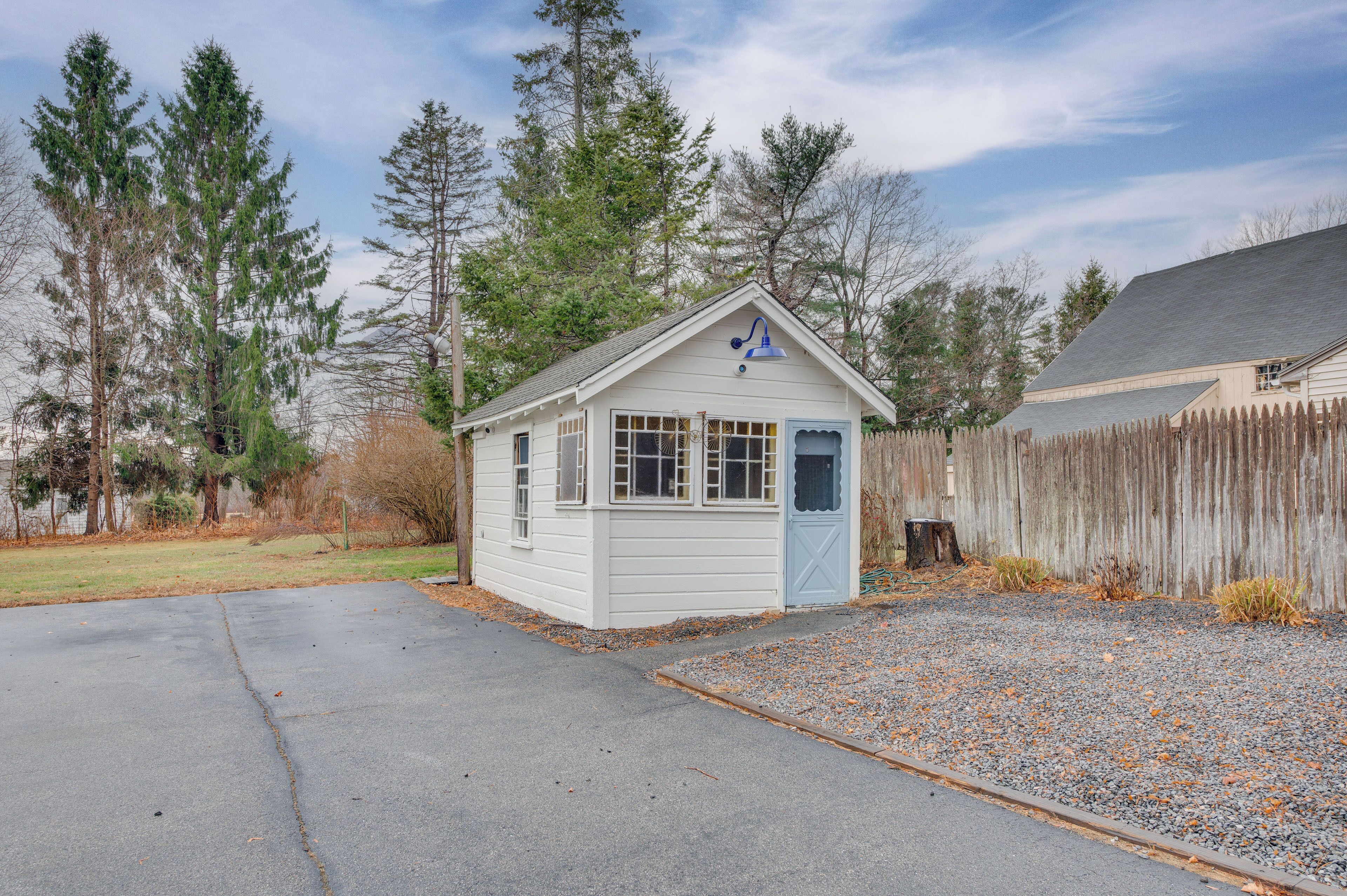 A small, single-story shed with a gabled roof and a blue door, surrounded by a wooden fence and trees.