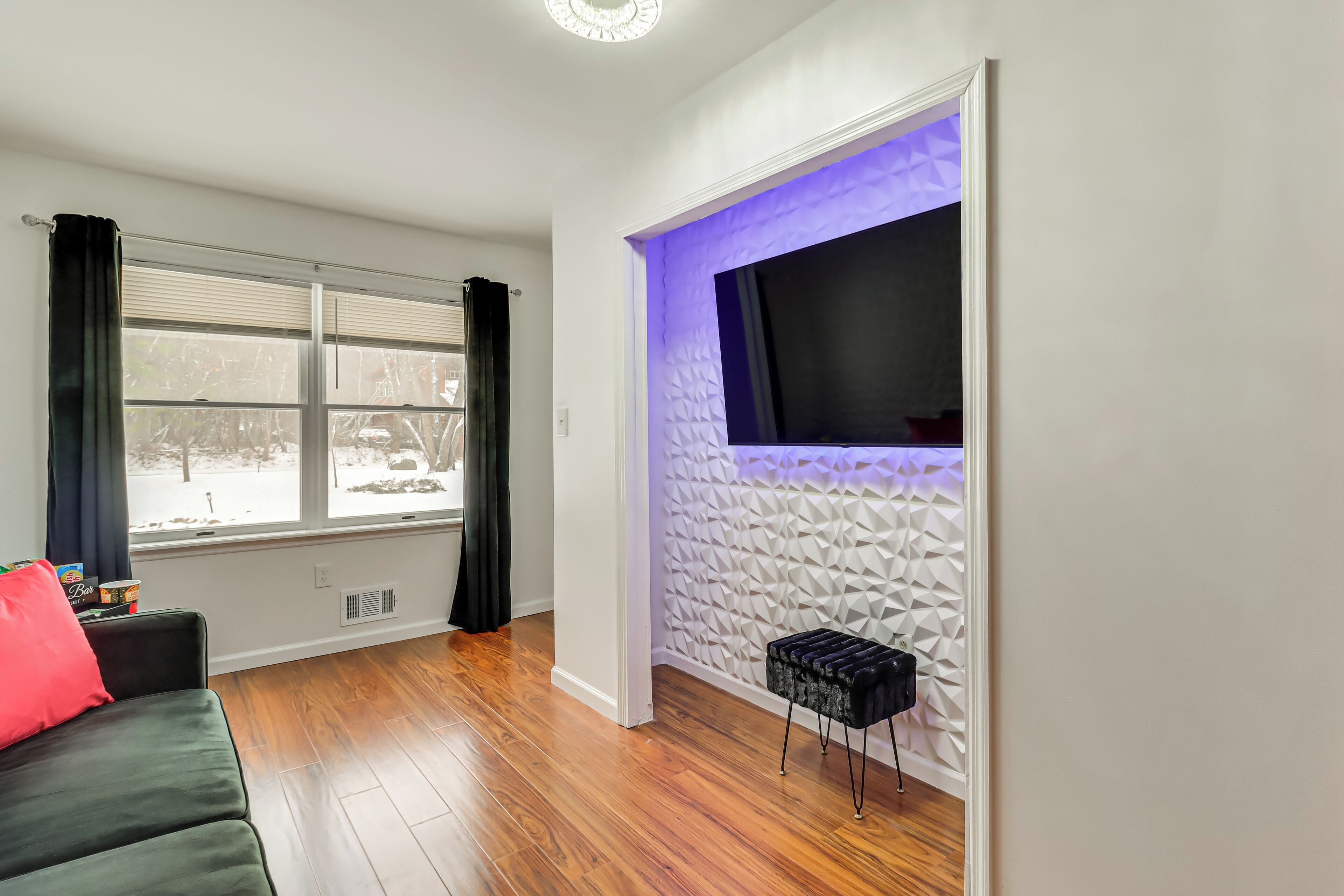 A modern living room with a large window, a black sofa, a small black ottoman, and a textured wall with a recessed TV.