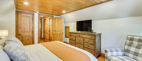 Quarto com teto de madeira, cama com manta bege, cômoda de madeira, televisão em cima e poltrona xadrez.