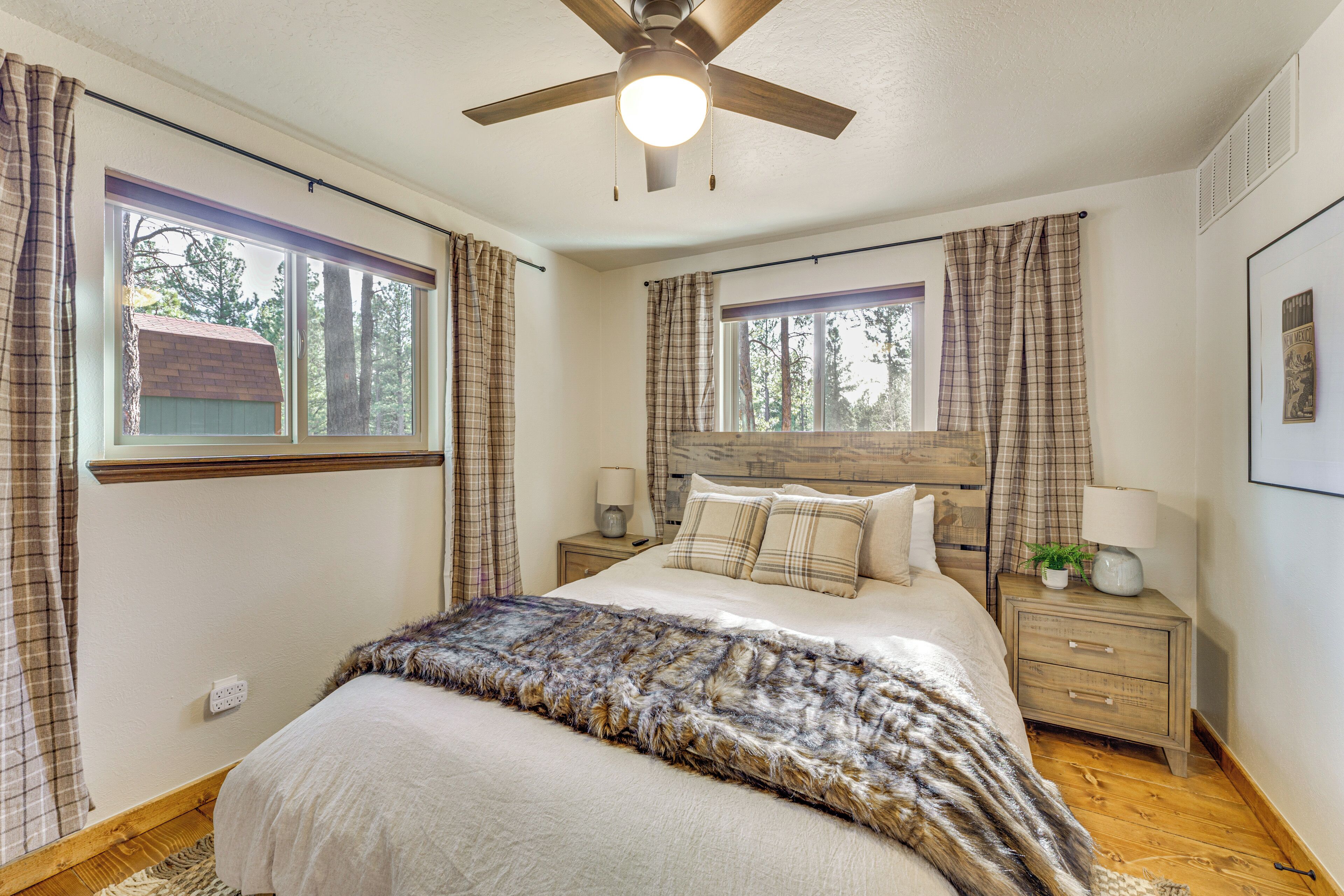 A bedroom with a bed, a ceiling fan, two bedside tables with lamps, plaid curtains, and a window with a view of trees.