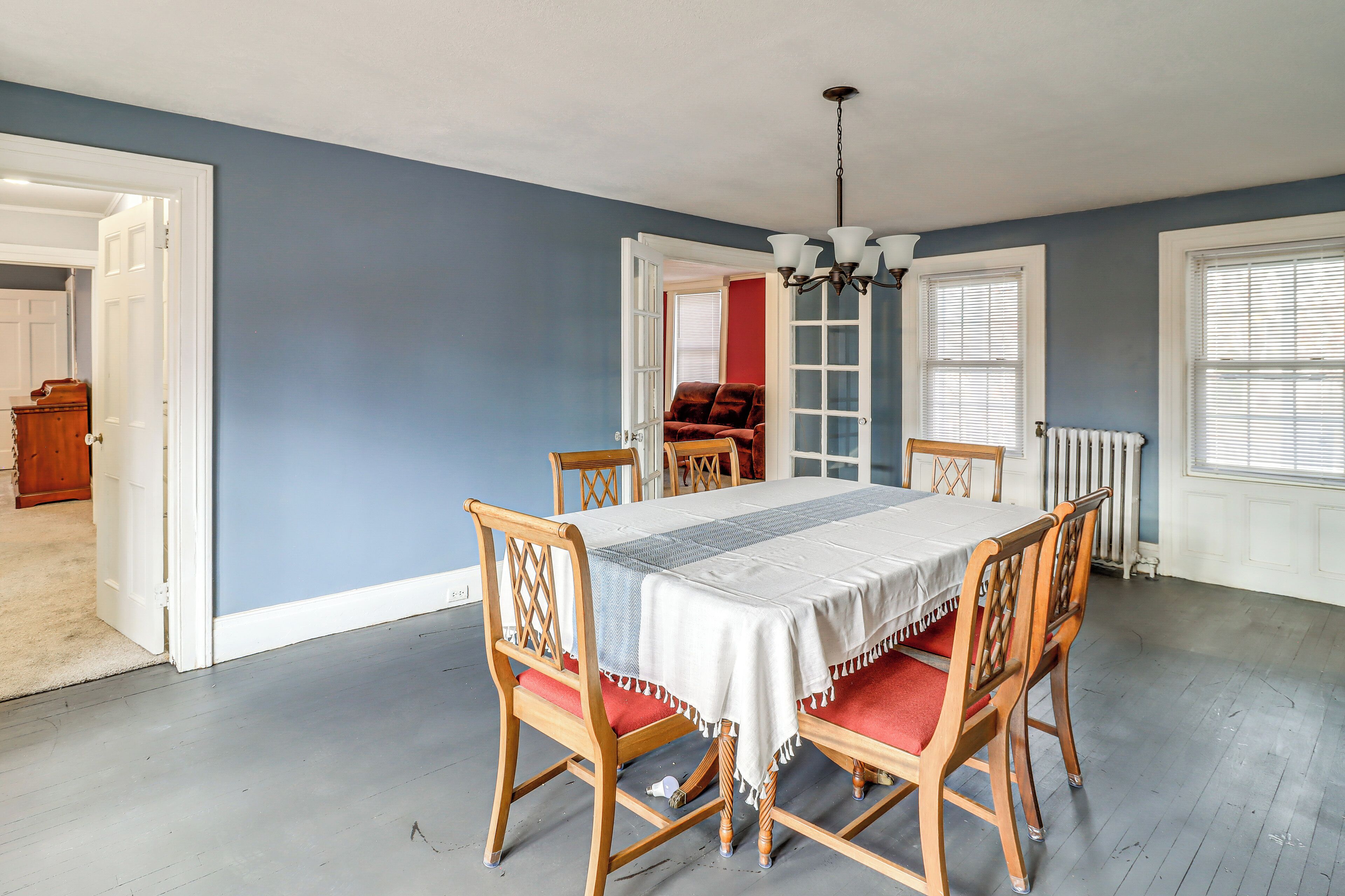 A dining room with a table set for four, wooden chairs with red cushions, a chandelier, and a view through an open door to another room.