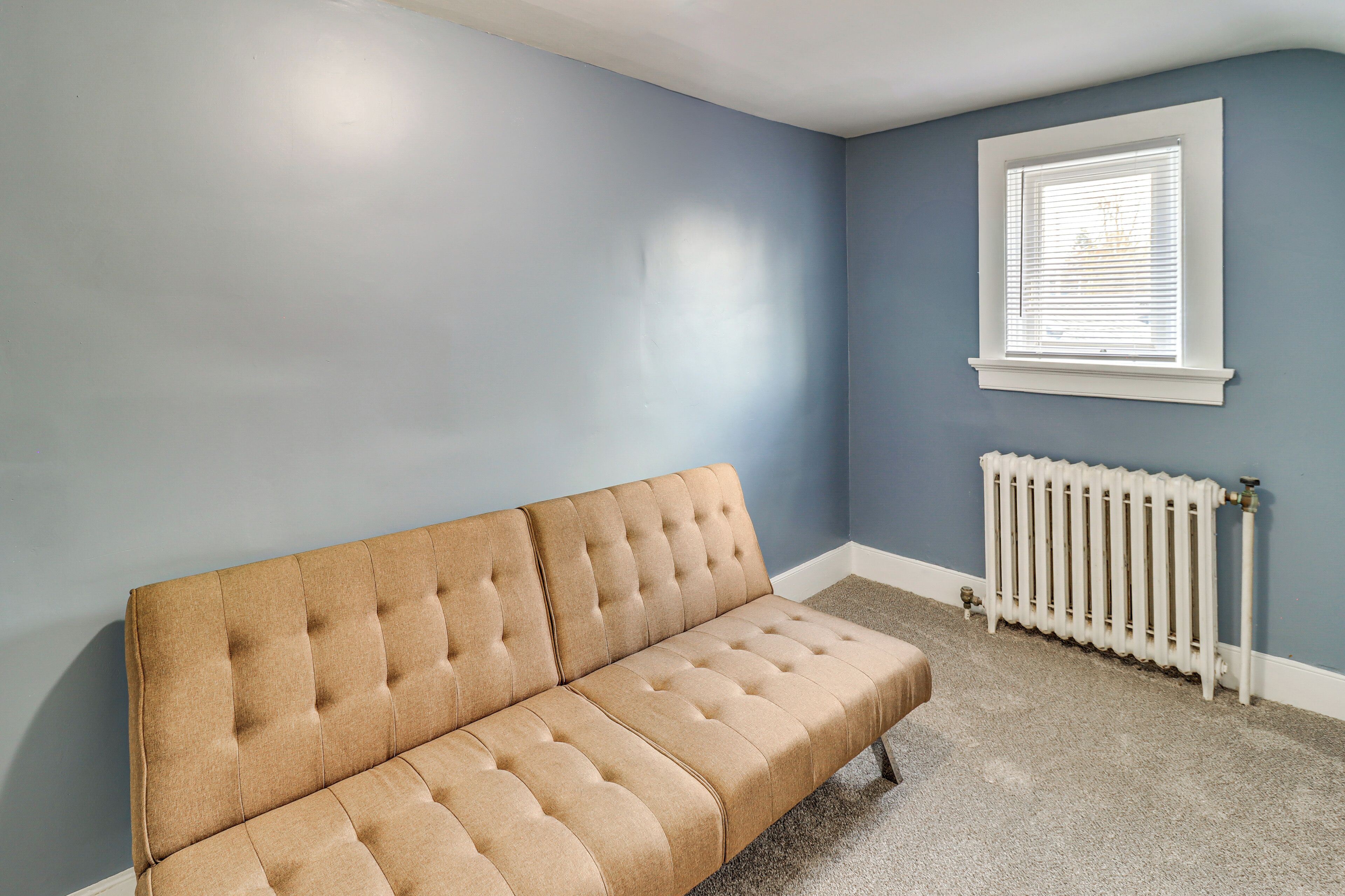 A room with a beige tufted sofa, a white radiator, and a window with blinds.