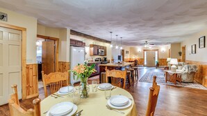 Un coin repas avec une table ronde dressée pour quatre personnes, des chaises en bois, un vase avec des fleurs et, au fond, une cuisine.