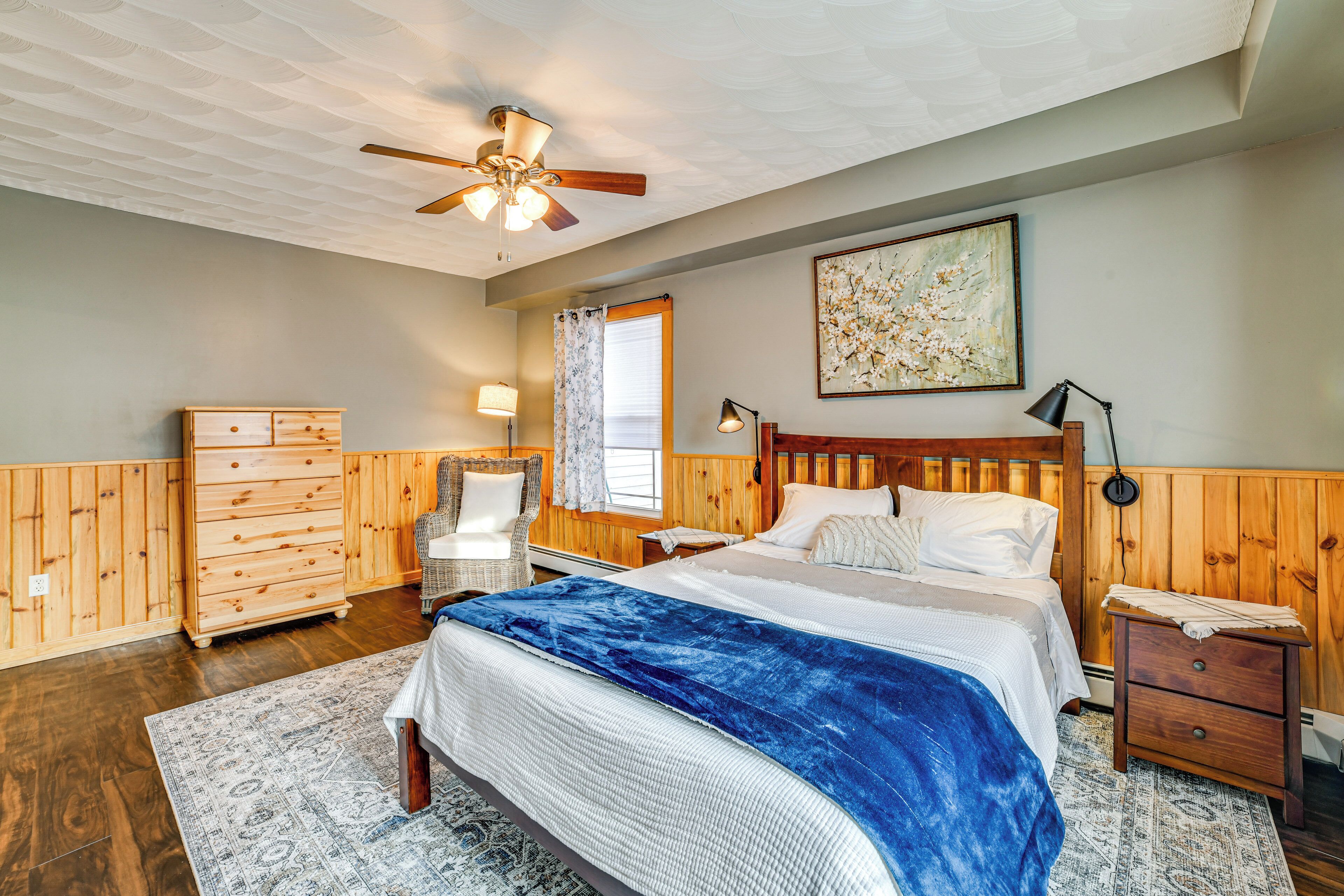 A bedroom with a bed, wooden headboard, nightstands, a dresser, a chair, a ceiling fan, and a window with curtains.