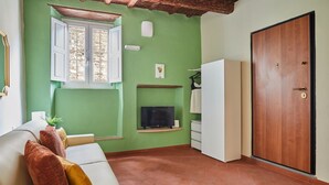 Une pièce avec un mur vert, une porte en bois, un canapé blanc avec des coussins, un téléviseur posé sur une étagère et une fenêtre dont les volets sont ouverts.