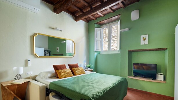 Une chambre avec un lit, une table de chevet avec une lampe, un miroir, une fenêtre avec des volets et un téléviseur posé sur une étagère.