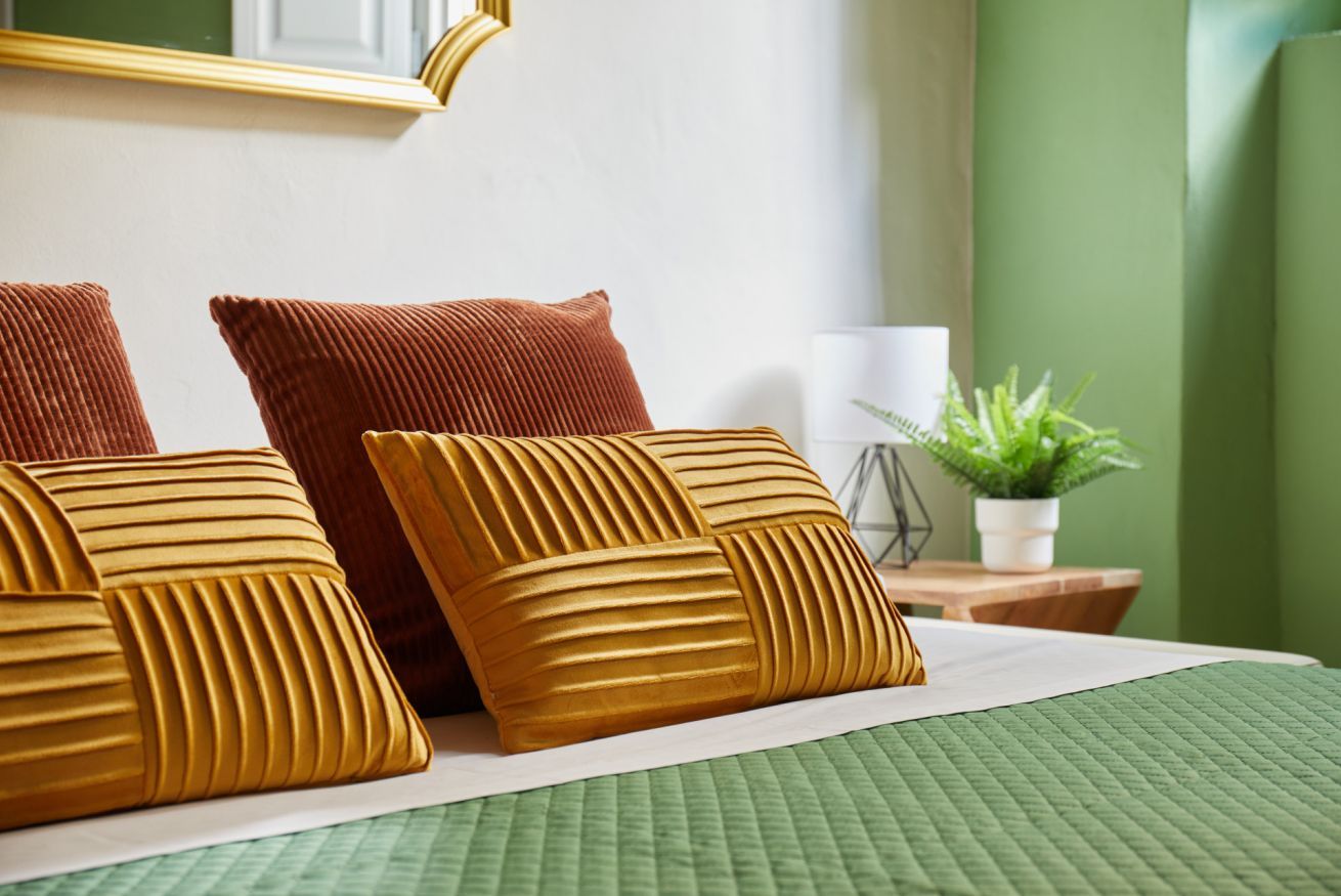 A neatly made bed with a green quilt, two brown pillows, and a small potted plant on a wooden nightstand.