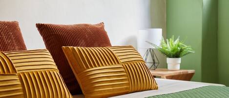 Una cama bien hecha con un edredón verde, dos almohadas marrones y una pequeña planta en maceta sobre una mesita de noche de madera.