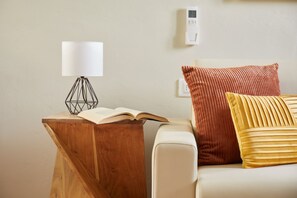 Een houten nachtkastje met een lamp, een open boek en een kussen op een witte bank.