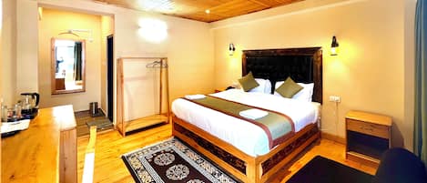 Une chambre d’hôtel avec un plafond en bois, un grand lit avec une tête de lit, des lampes de chevet, une table de chevet en bois, une petite table, un miroir, un espace douche et un tapis à motifs.