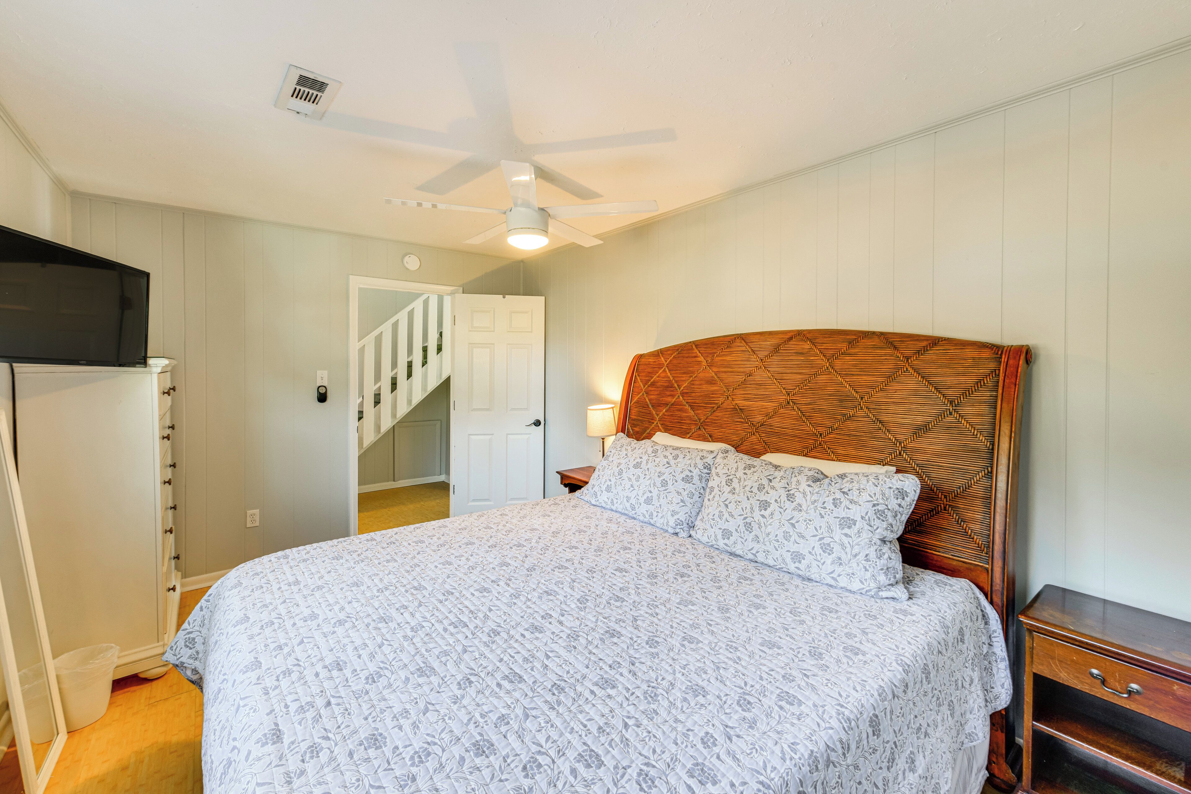 A bedroom with a bed, a nightstand, a wooden headboard, a ceiling fan, and a door leading to a staircase.