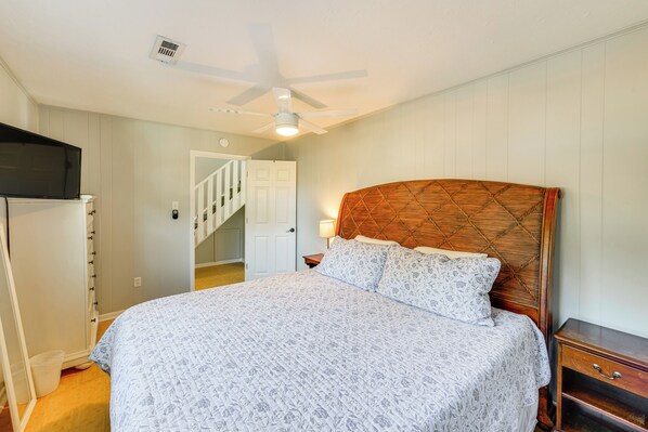 A bedroom with a bed, a nightstand, a wooden headboard, a ceiling fan, and a door leading to a staircase.