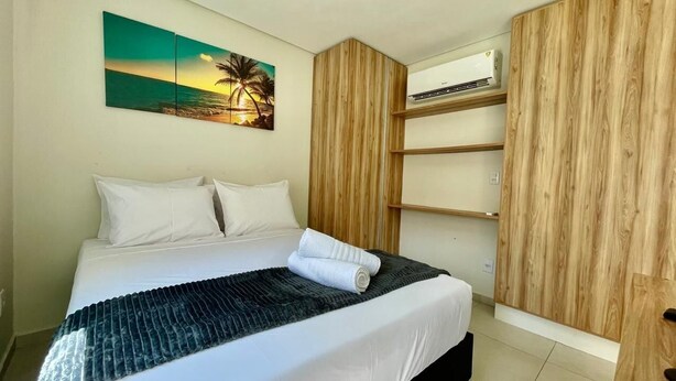 A bedroom with a bed, white pillows, a dark blanket, a wooden wardrobe, and a wall-mounted air conditioner.