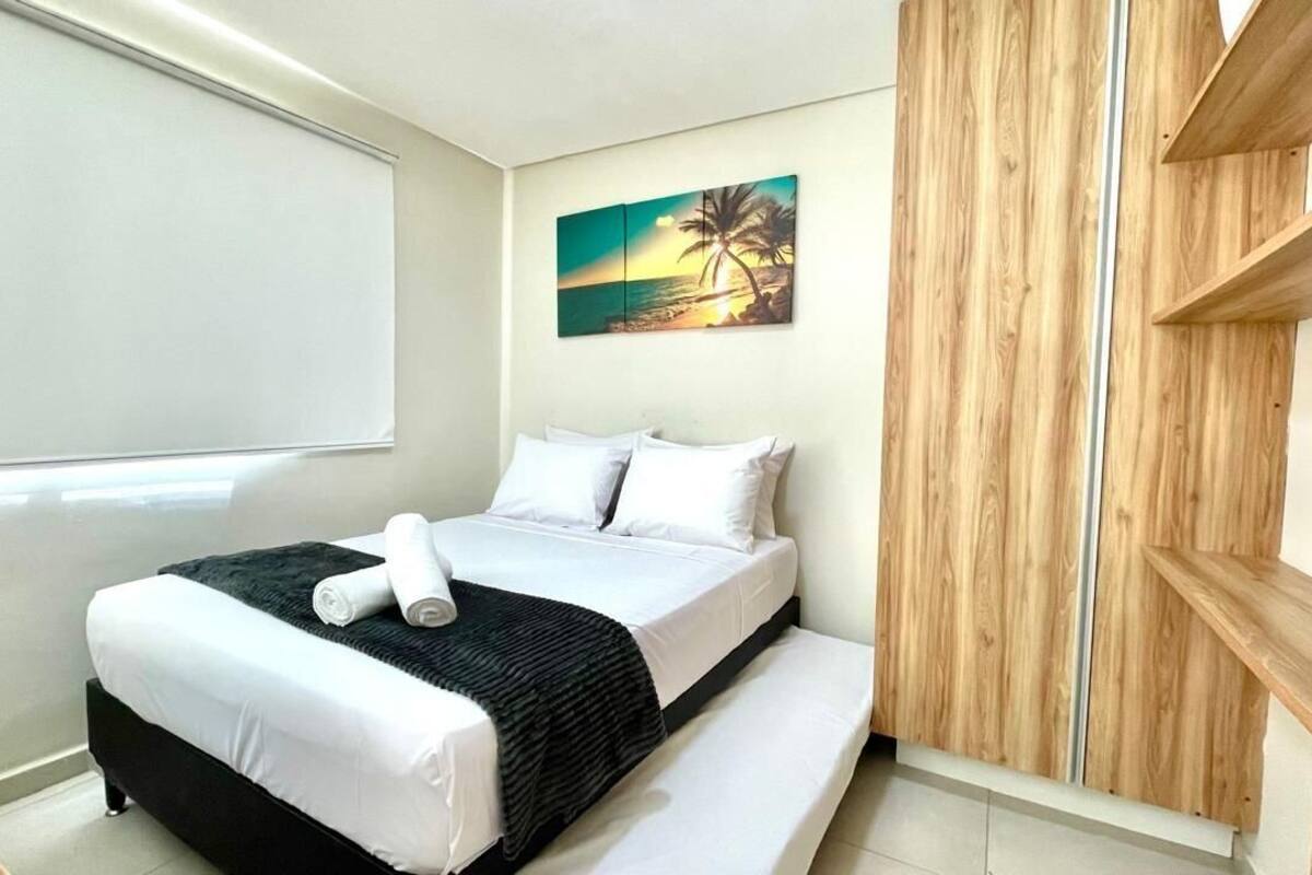 Un lit bien fait, avec du linge de lit blanc et un plaid noir, un tableau représentant un coucher de soleil accroché au mur, et un lambris en bois sur le côté droit.