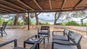 Un espace extérieur aménagé avec des tables et des chaises en bois, offrant une vue sur la mer et les montagnes, et surmonté d’une pergola.