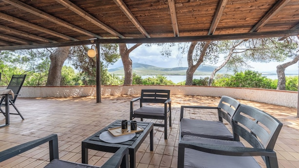 Un espace extérieur aménagé avec des tables et des chaises en bois, offrant une vue sur la mer et les montagnes, et surmonté d’une pergola.