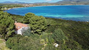An aerial view of a coastal property with a red-roofed building surrounded by dense greenery, overlooking a clear blue sea and a distant shoreline with buildings. - Villa Laura by Wonderful Italy (Alghero)