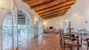 Une salle à manger spacieuse avec un plafond haut et un sol carrelé, comprenant une table et des chaises en bois, ainsi qu’un téléviseur posé sur un meuble.